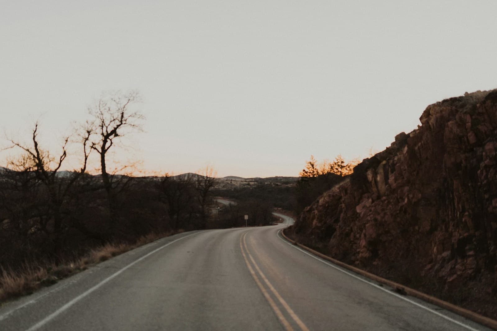 Empty road winding through desert landscape at sunset with rocky cliffs and bare trees