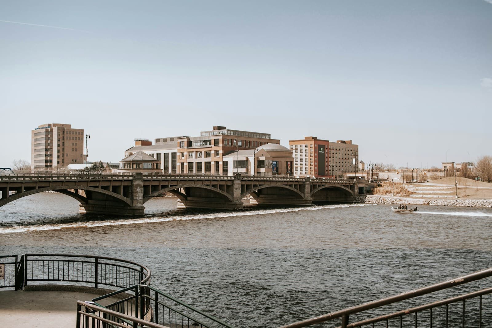 Stone arch bridge over river with urban buildings and overcast sky, viewed from waterfront promenade