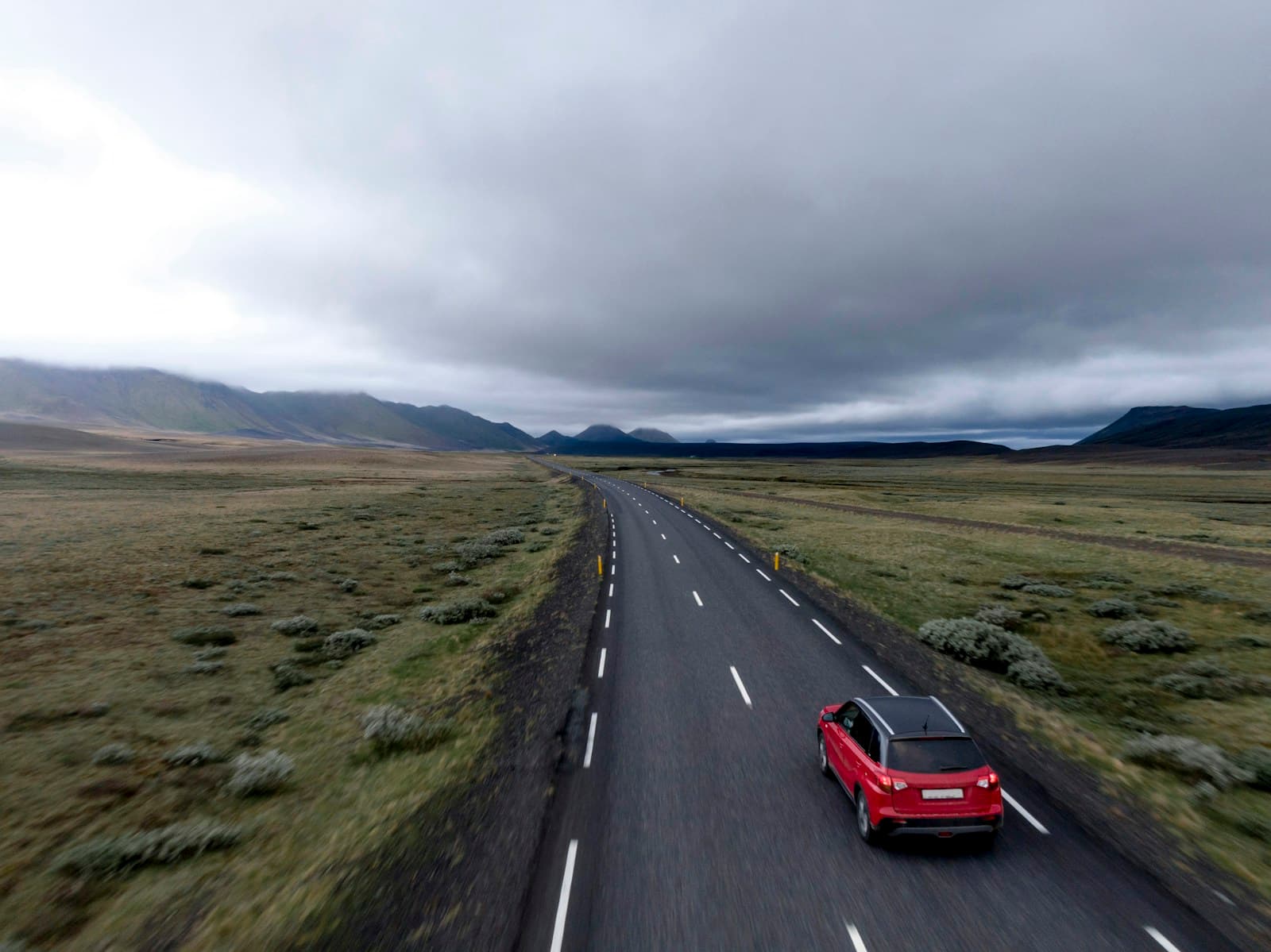 Red car driving on empty highway through remote landscape with mountains and cloudy sky