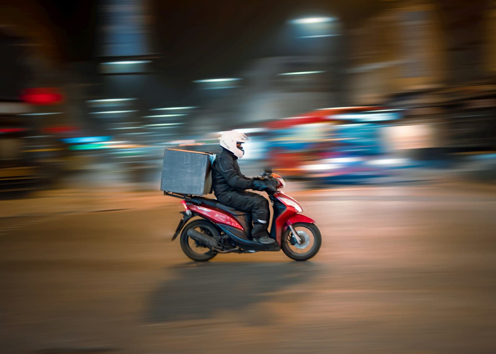 Delivery rider on a red scooter speeding through a busy city street at night