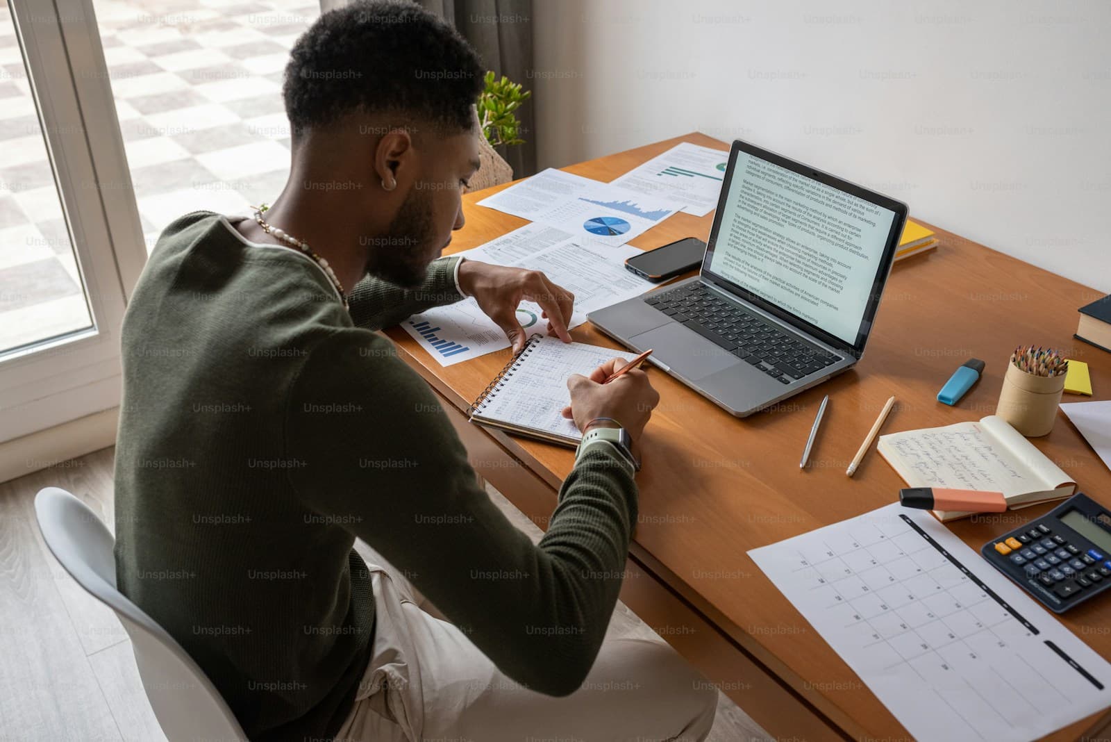 Young man writing on papers at a desk by a window with a laptop open