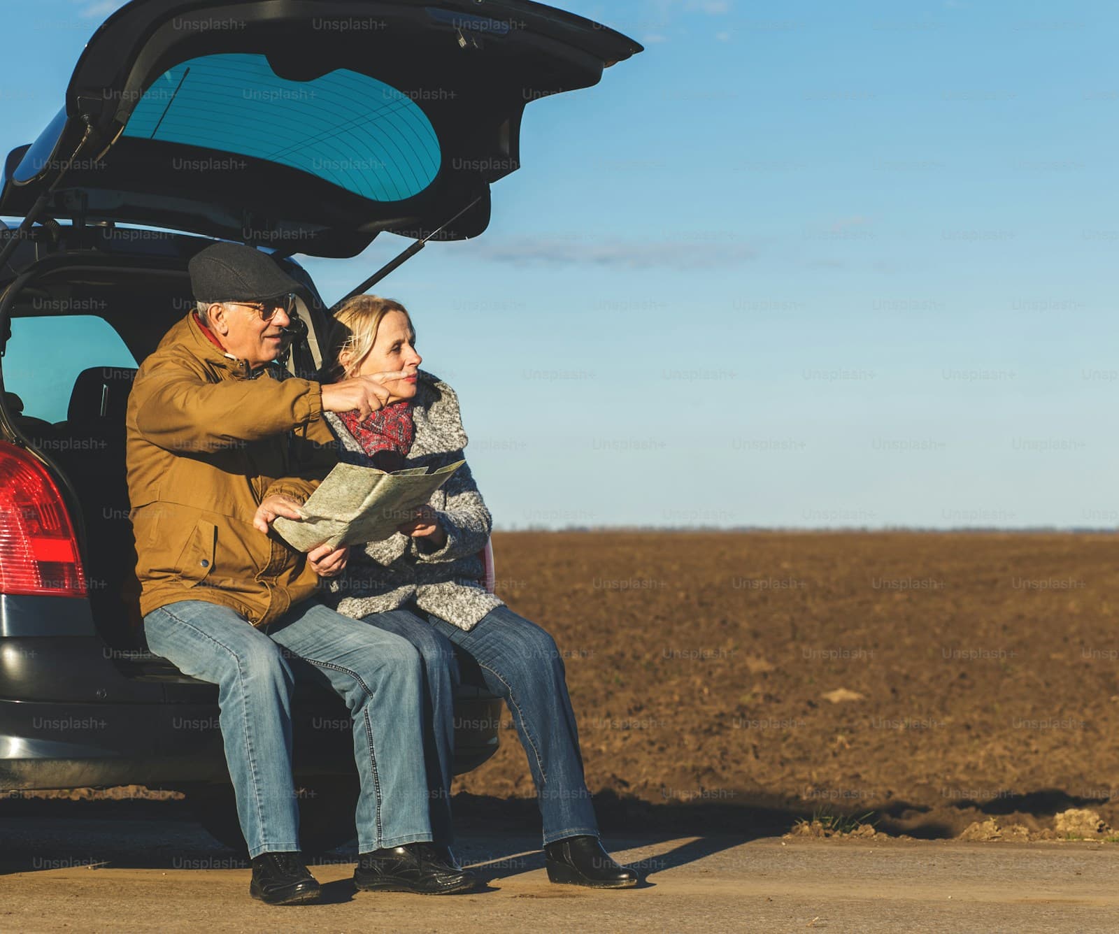 Elderly couple resting together on the open tailgate of their SUV in a rural setting