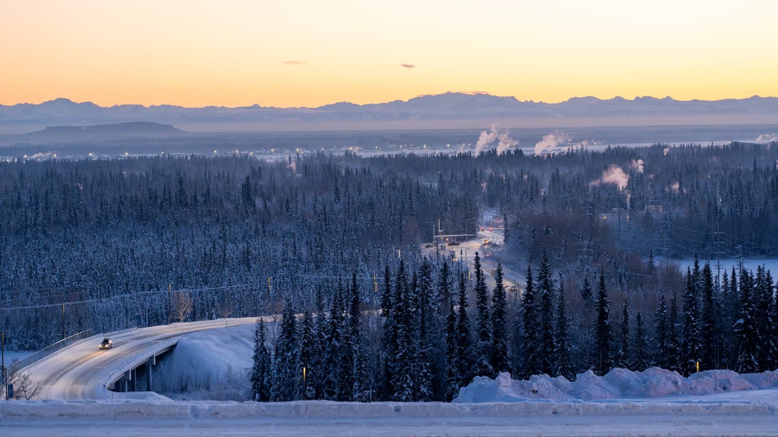 Snowy road through evergreen forest at sunset with mountains in background during winter
