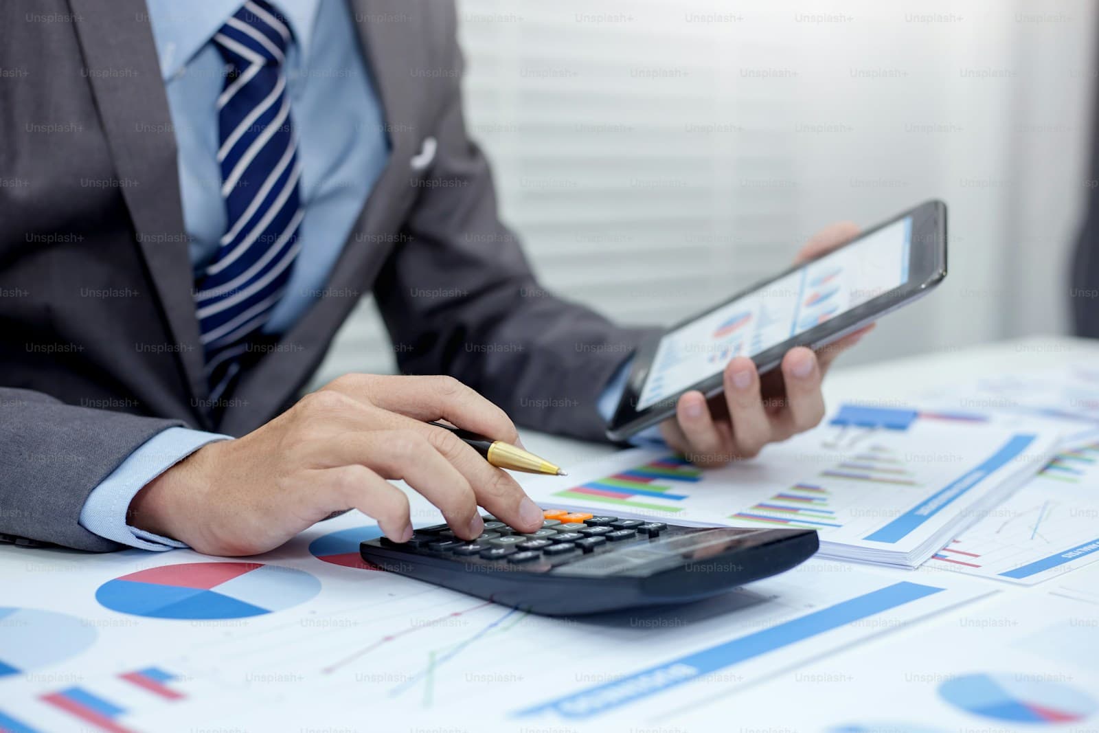 Person in a gray suit using a calculator and tablet among printed charts