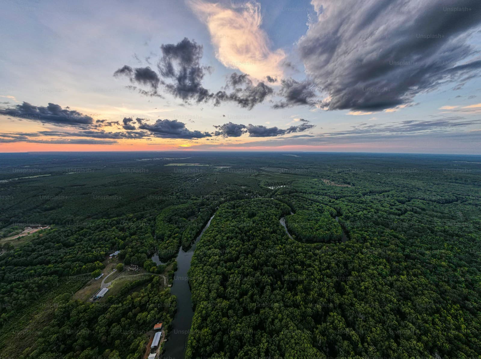 Aerial view of dense forest with winding river at sunset, dramatic clouds in colorful sky over treetops