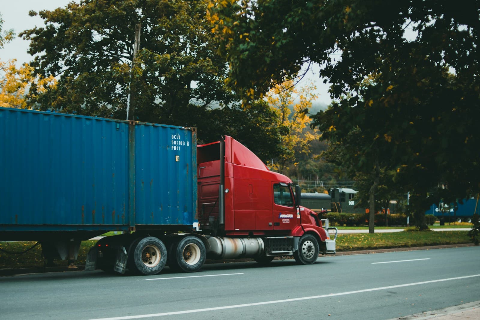 Red semi-truck hauling a blue shipping container down a tree-lined road at dusk