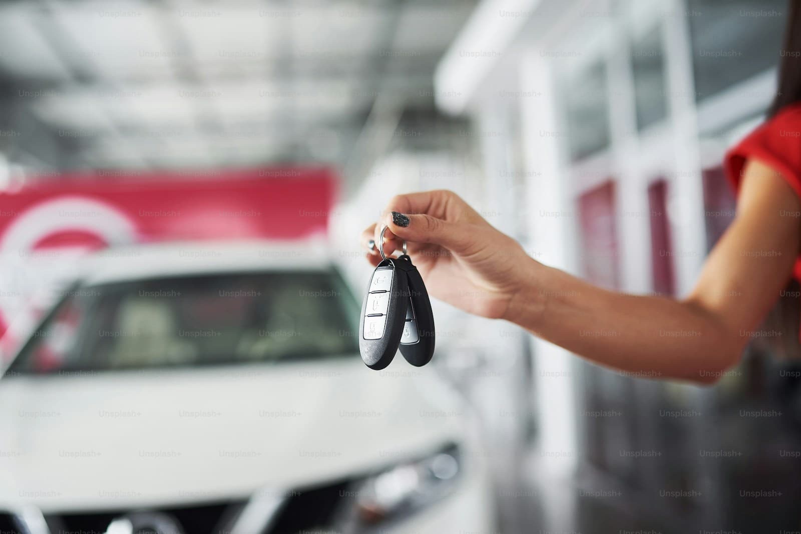 Hand holding black car keys with white car and red dealership signage blurred in background