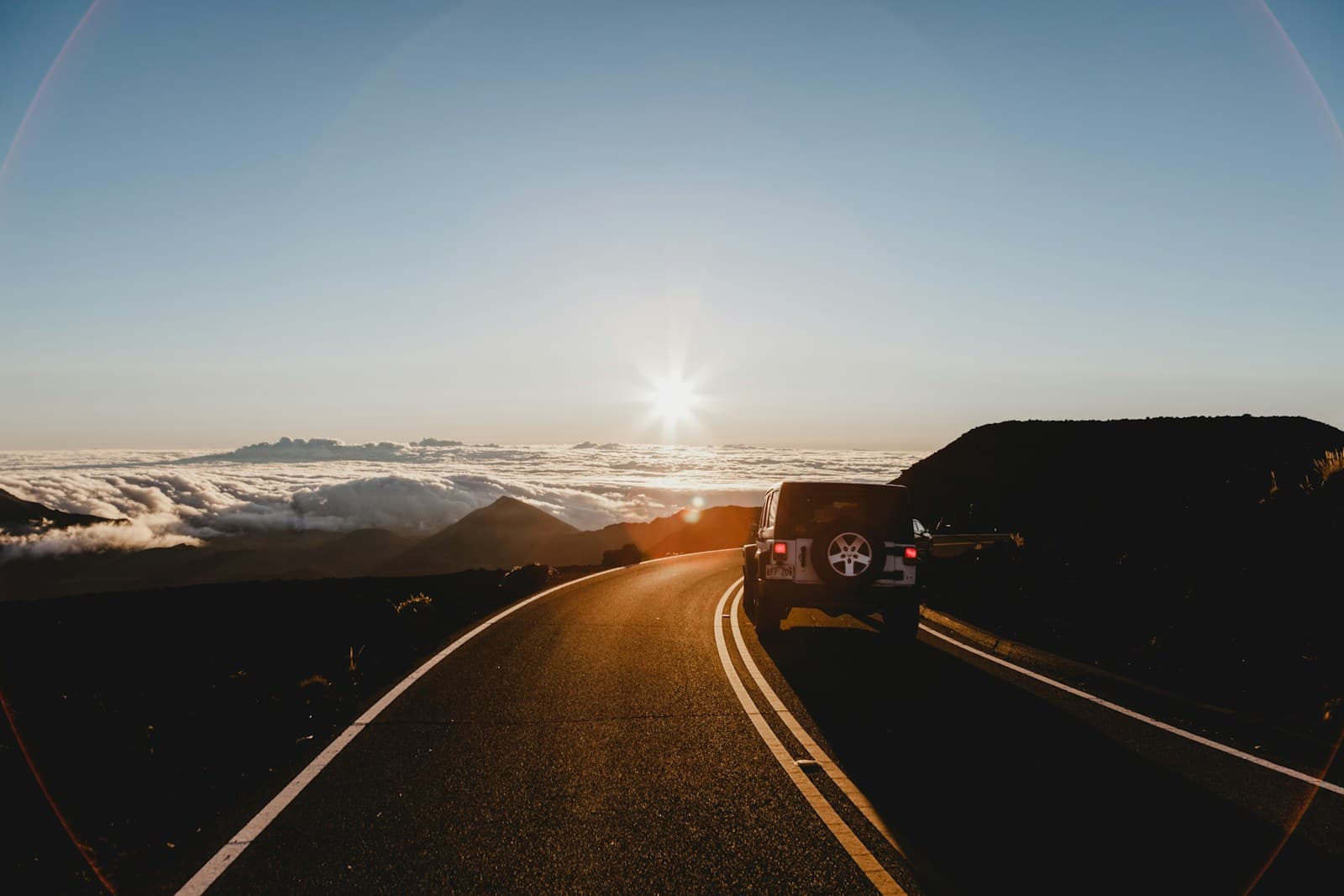 Mountain road at sunset with car driving toward bright sun, clouds below in valley, golden hour lighting