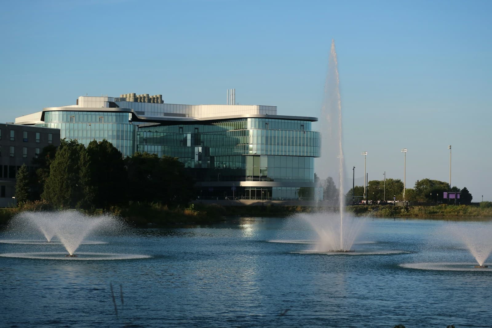 Modern glass building on waterfront with multiple water fountains spraying in lake under clear blue sky