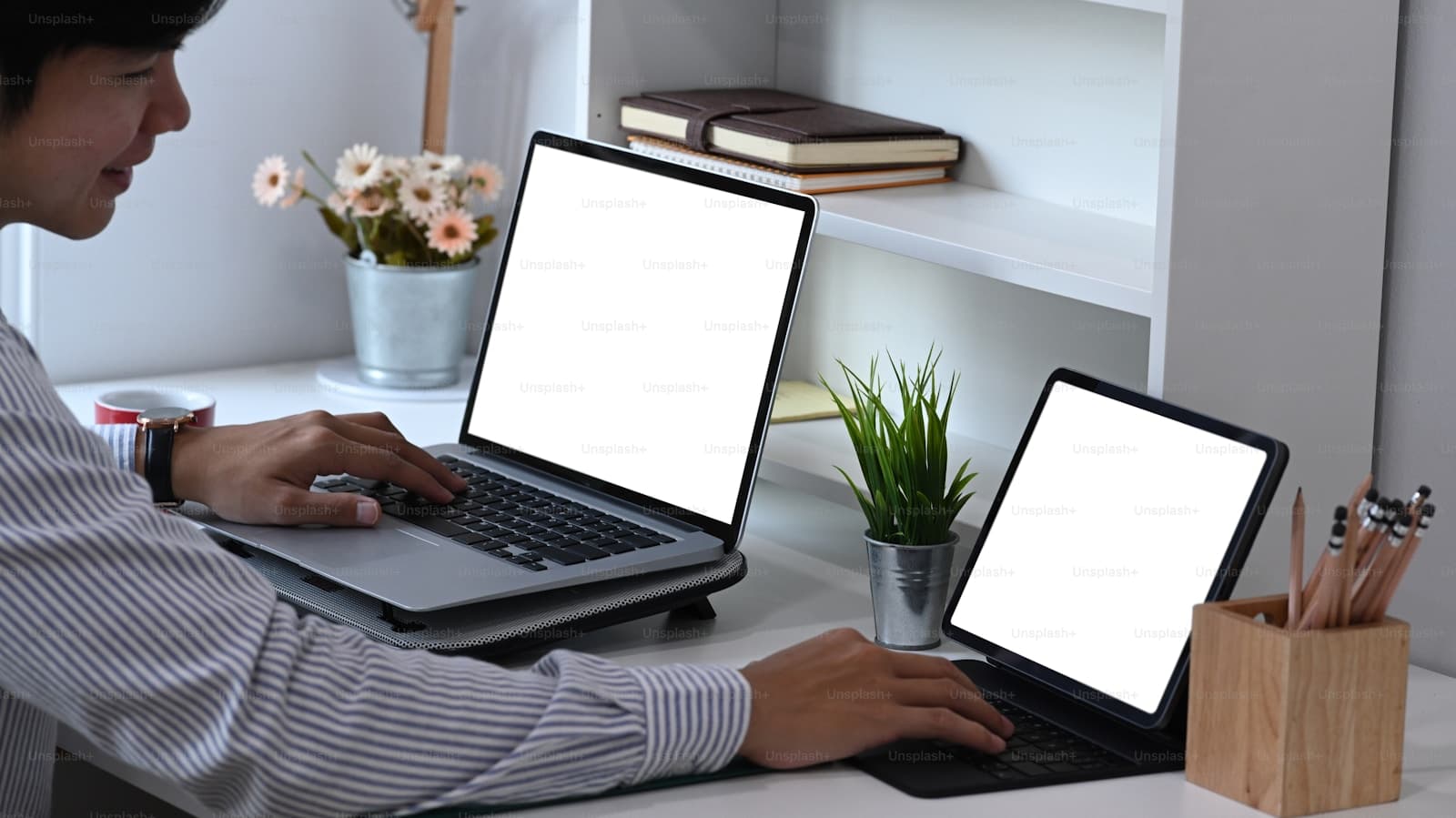 Person in a striped shirt typing at a white desk with two open laptops