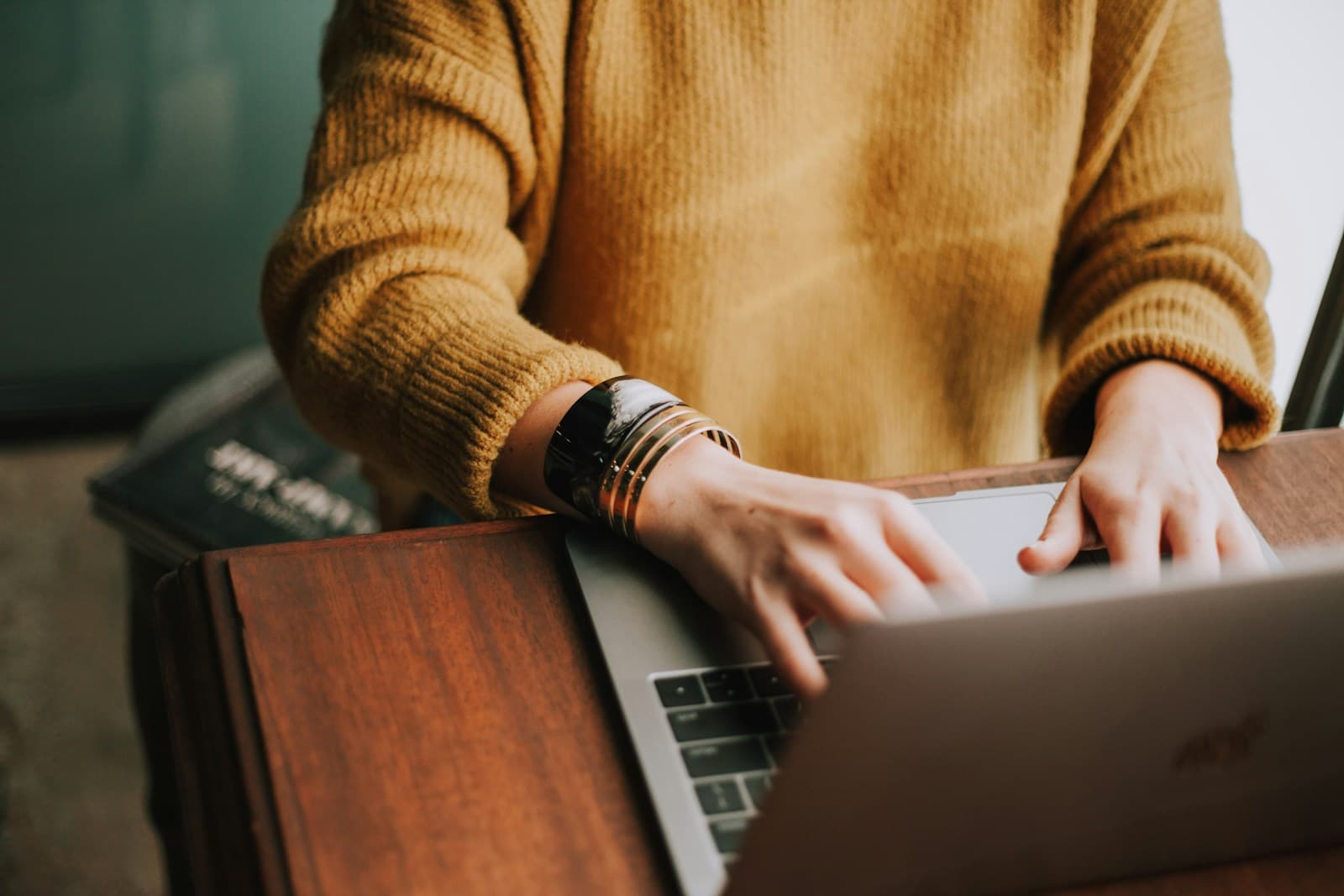 Person in a mustard knit sweater typing on a silver laptop at a wooden desk