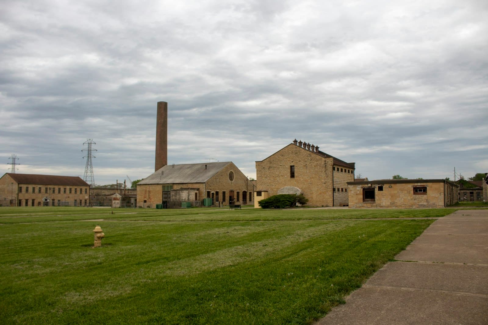 Historic brick industrial complex with smokestack and multiple buildings surrounded by green lawn under cloudy sky