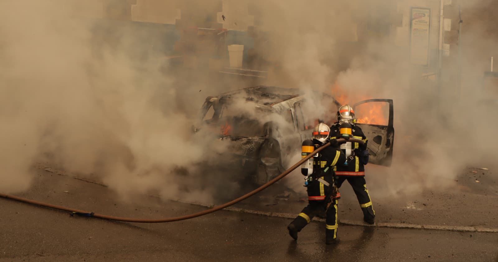Firefighters in protective gear using hoses to extinguish a vehicle fire with heavy smoke