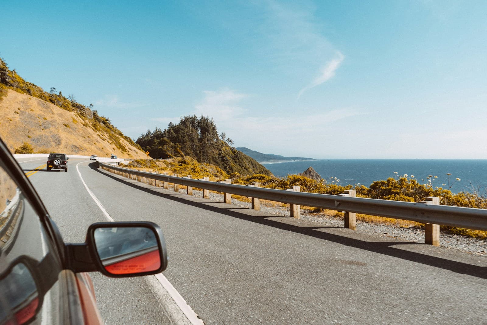 Curved coastal highway beside a blue ocean, seen past a car's side mirror