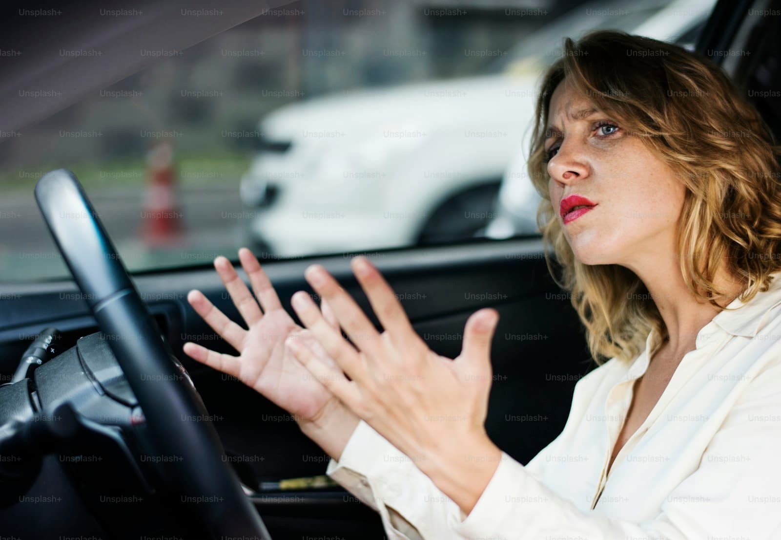Frustrated woman in a white blazer throwing both hands up from the steering wheel