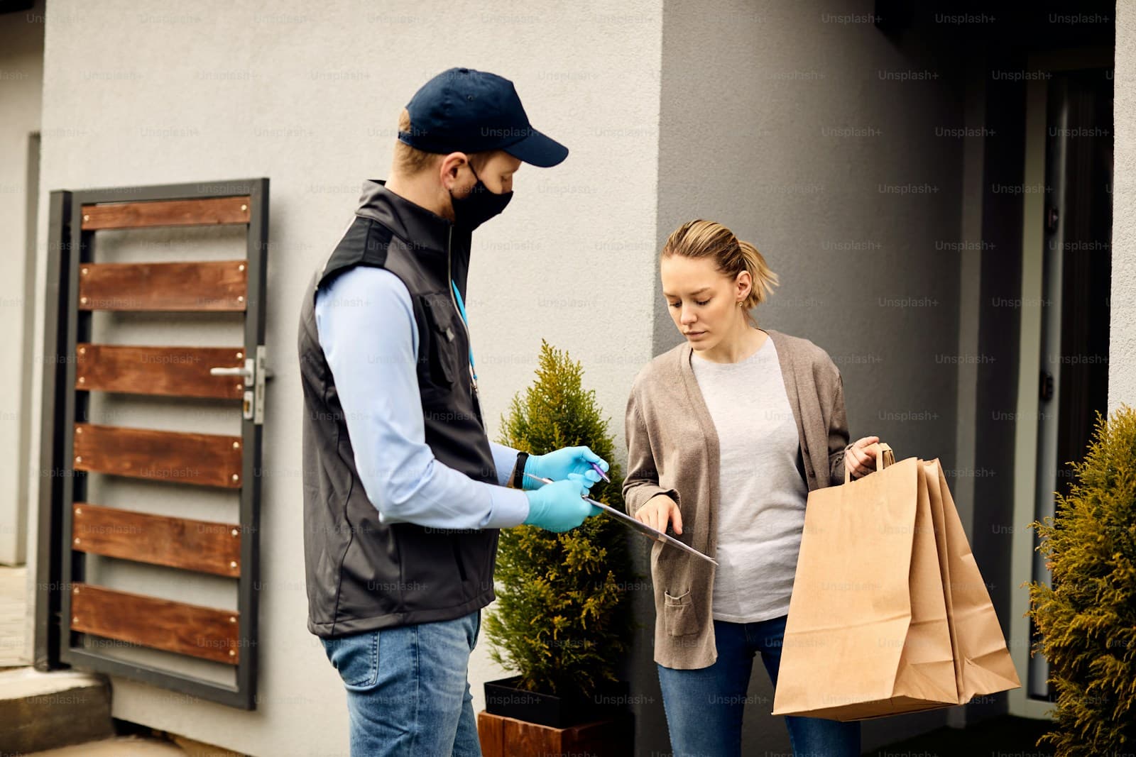 Delivery worker in a cap and vest handing a clipboard to a woman at her front door