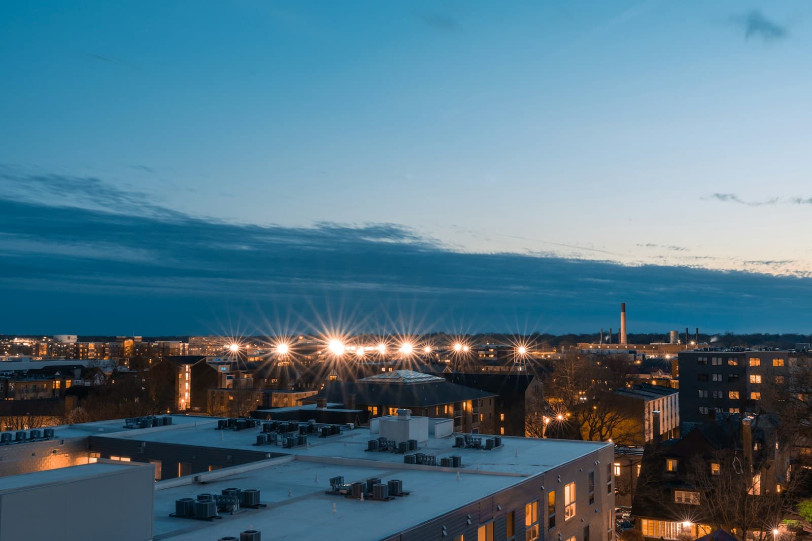 Winter twilight cityscape with snow-covered buildings and warm street lights creating starbursts against blue evening sky